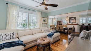 A spacious living room with a sectional sofa, ottoman, wooden dining table, and a ceiling fan.