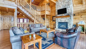 Interno di una baita di legno con camino in pietra, scala in legno e un accogliente soggiorno con divani e tavolino.