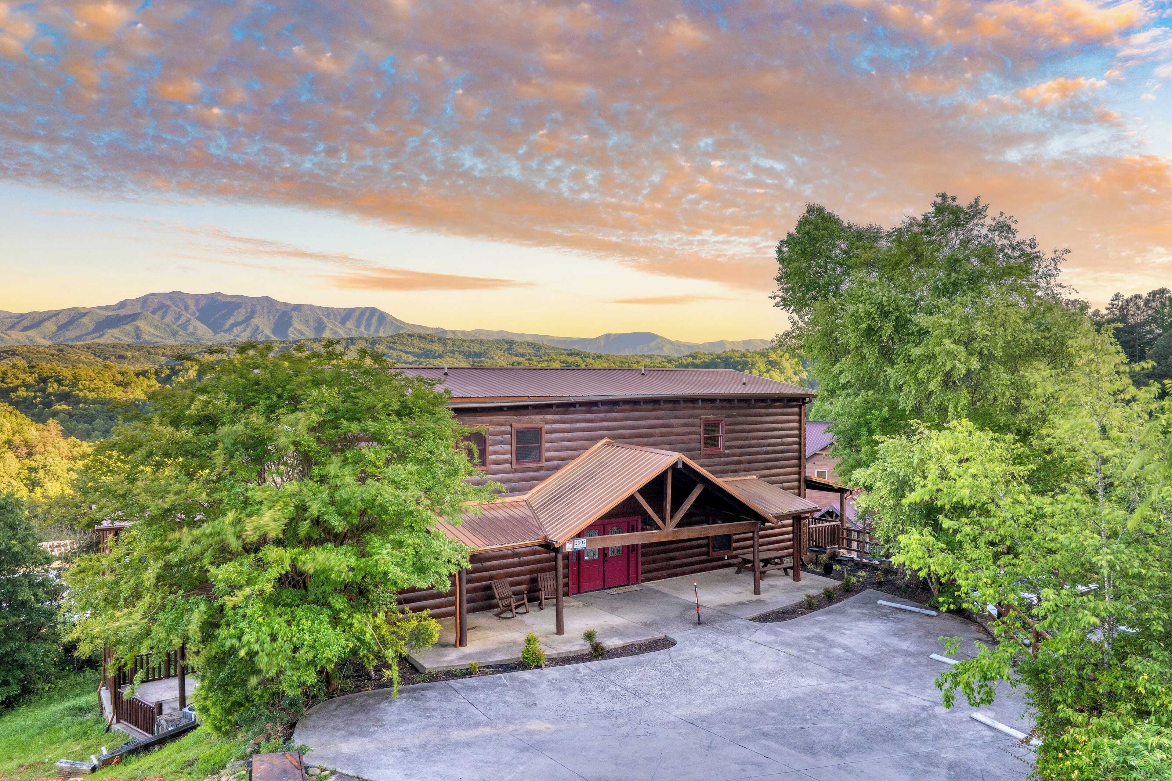 A rustic log cabin with a covered patio, surrounded by lush greenery and mountains in the background.