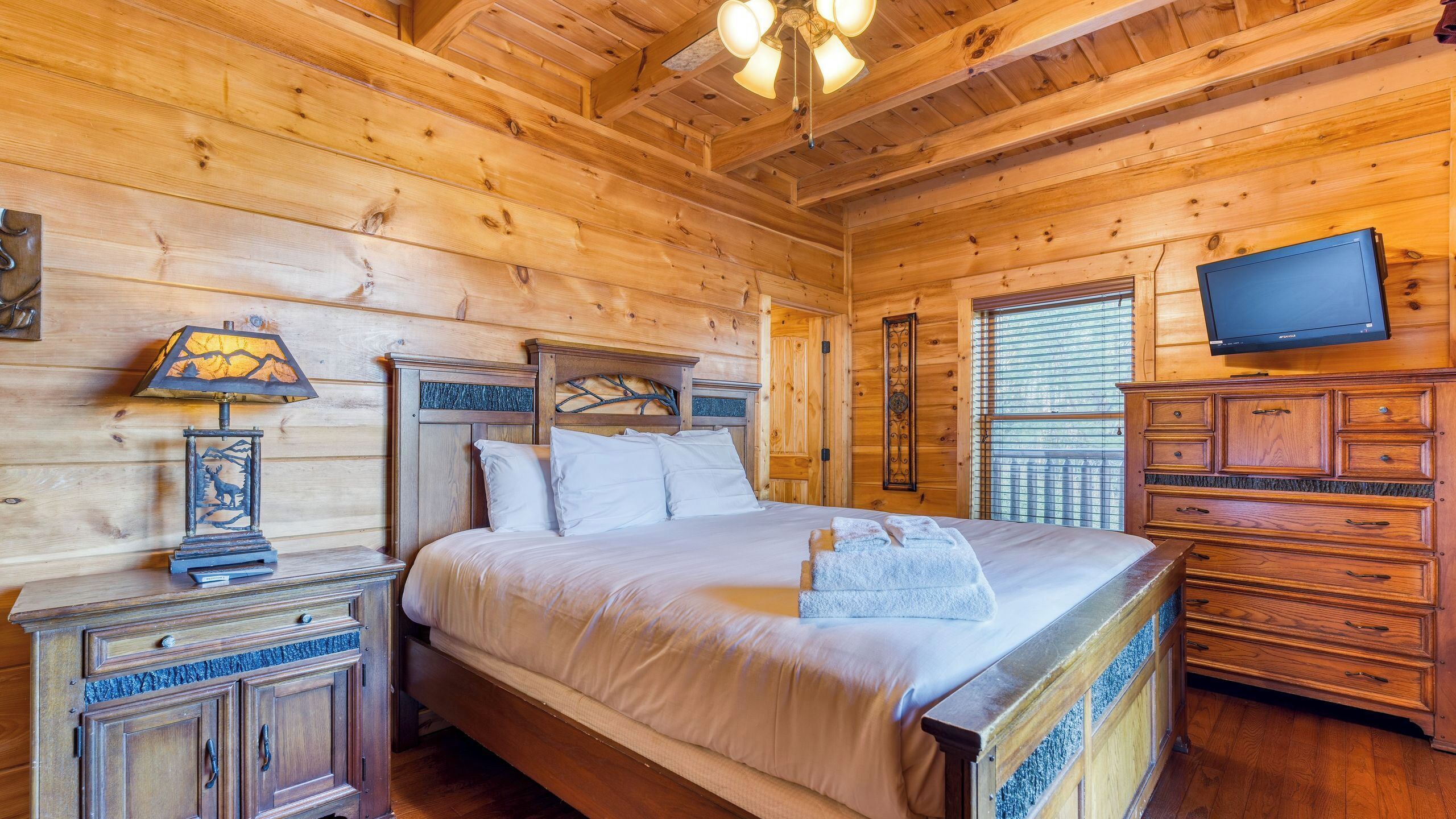 A wooden cabin bedroom with a bed, a nightstand with a lamp, a dresser, and a television.