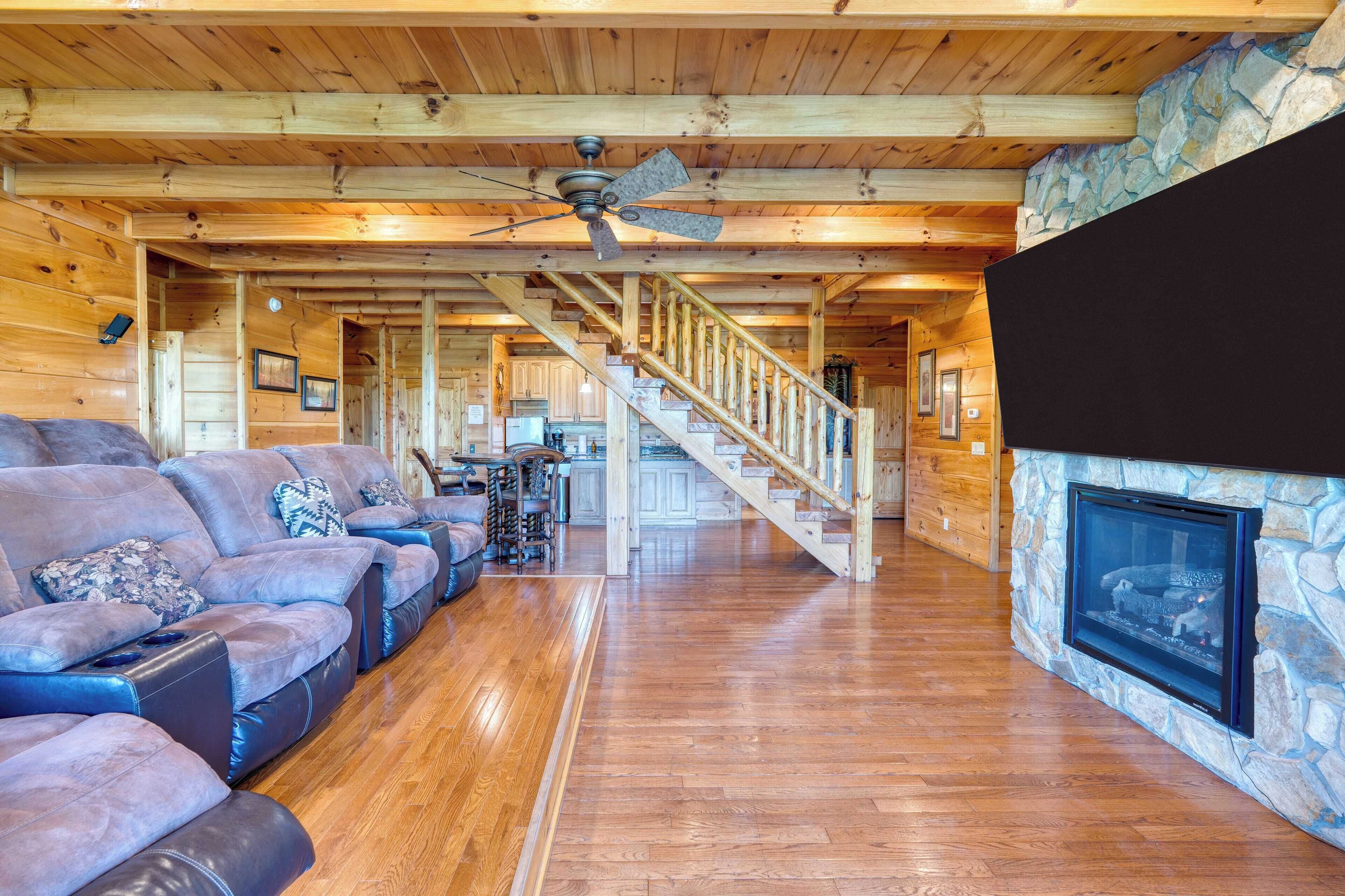A spacious living room with a stone fireplace, wooden ceiling, and a staircase leading to upper floors.