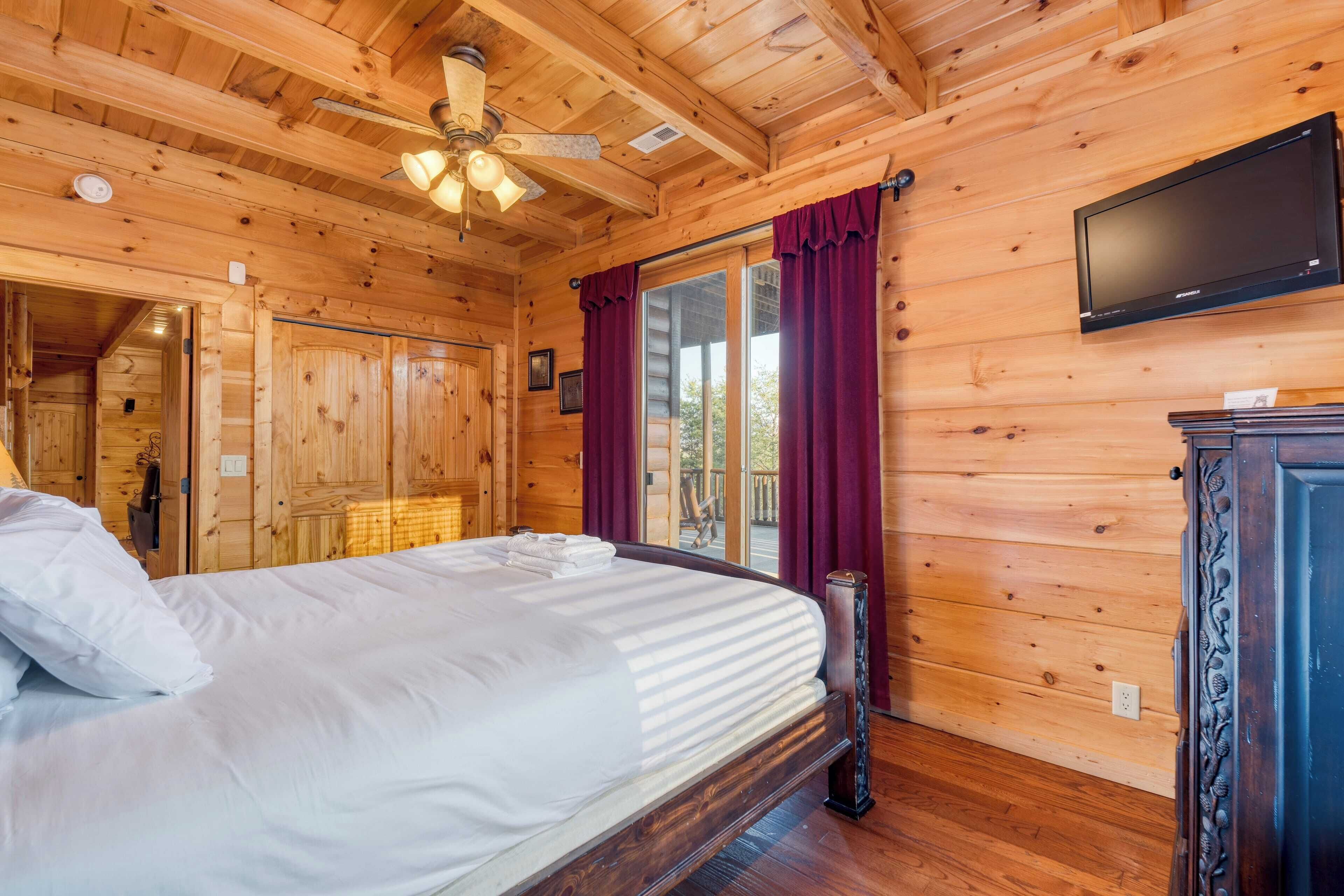 Ein Zimmer in einer Holzhütte mit einem Bett, einem Deckenventilator, einem Fernseher und einer Glastür mit Vorhängen.