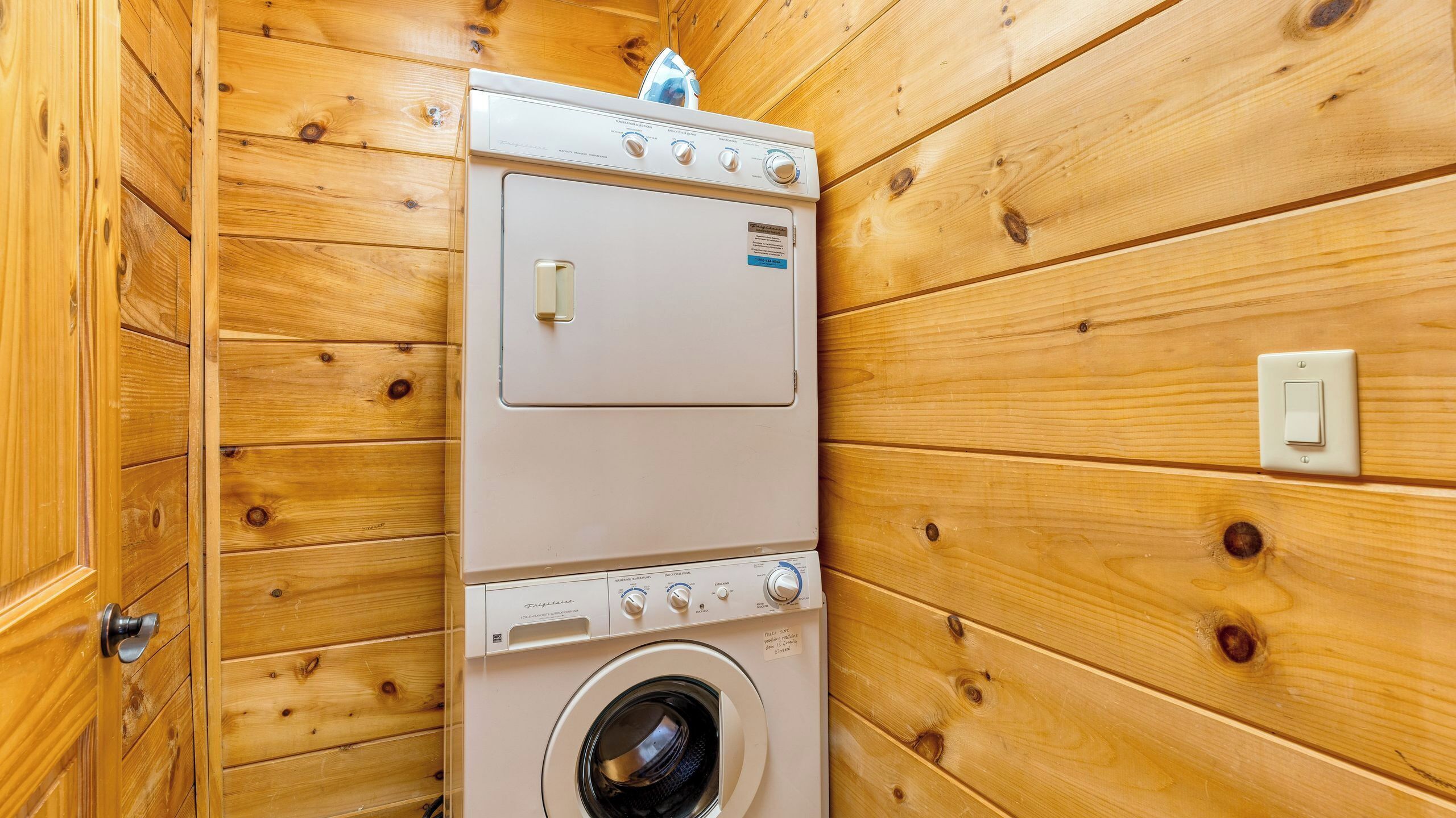 A compact, wooden-walled laundry room with a front-loading washer and dryer, a visible control panel, and a light switch on the wall.
