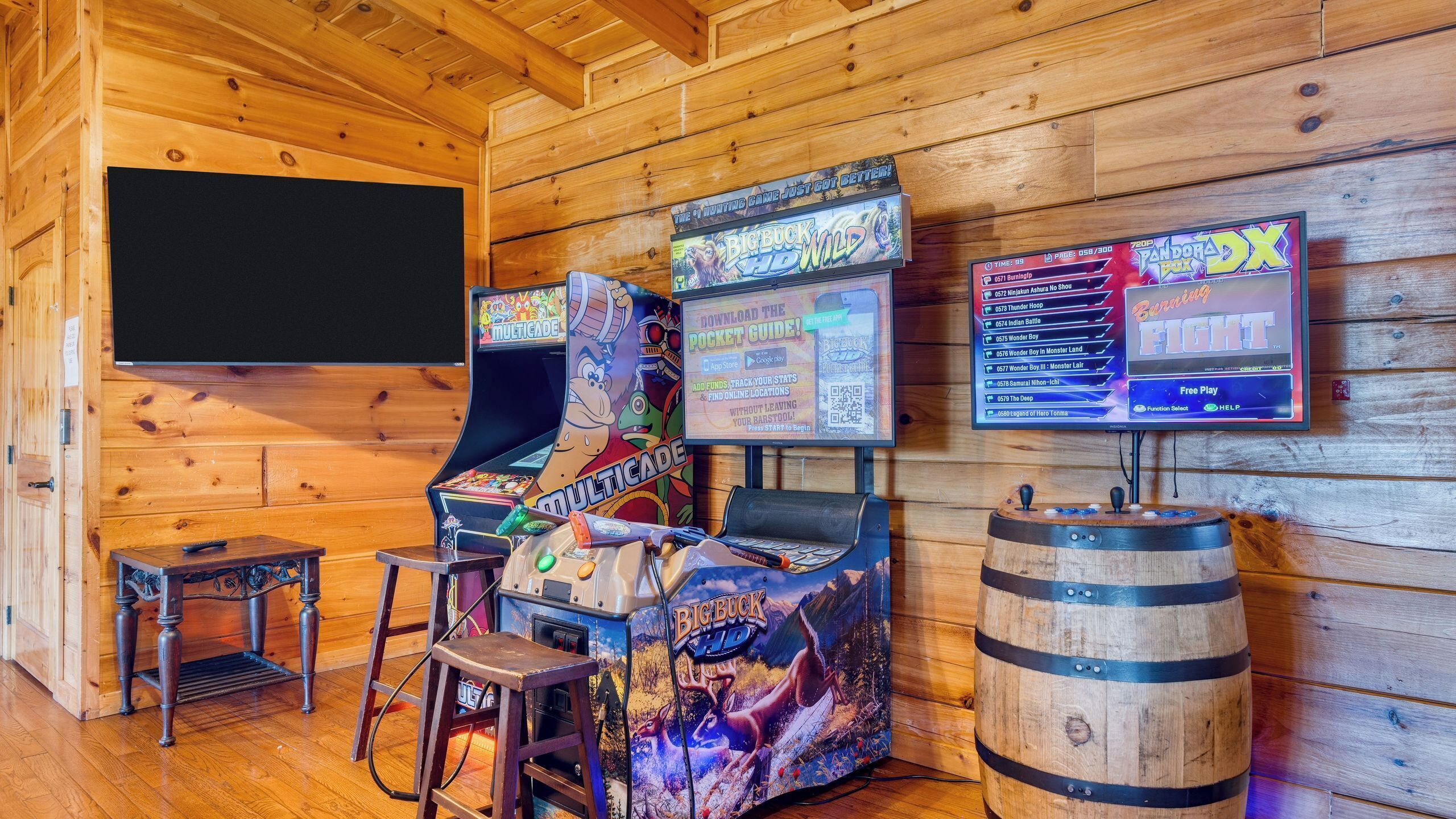 A wooden cabin with a gaming area featuring a Multicade arcade machine, a monitor displaying a game, and a wooden barrel.