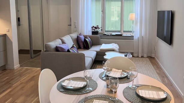 En moderne stue med sofa, spisebord dækket til fire personer og et stort vindue med gardiner.