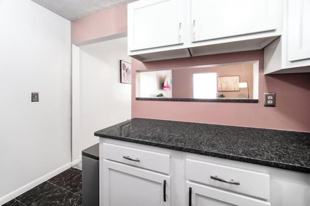 A kitchen with white cabinets, a black countertop, and a pink accent wall.