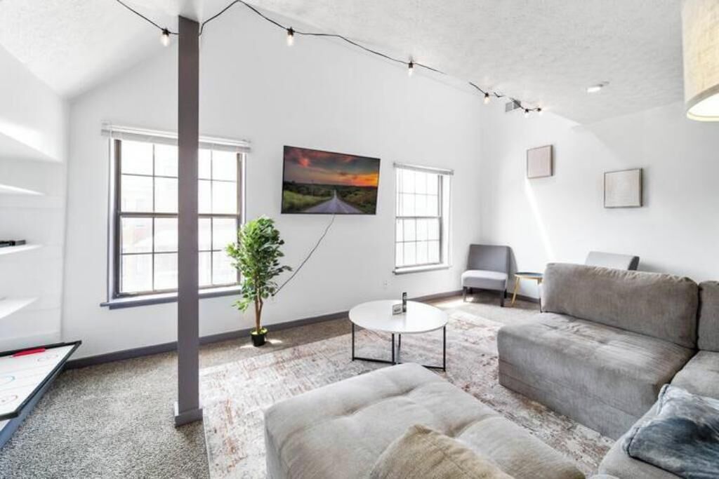 A modern living room with a large sofa, a round coffee table, and a painting on the wall. There are two windows and a plant in the room.