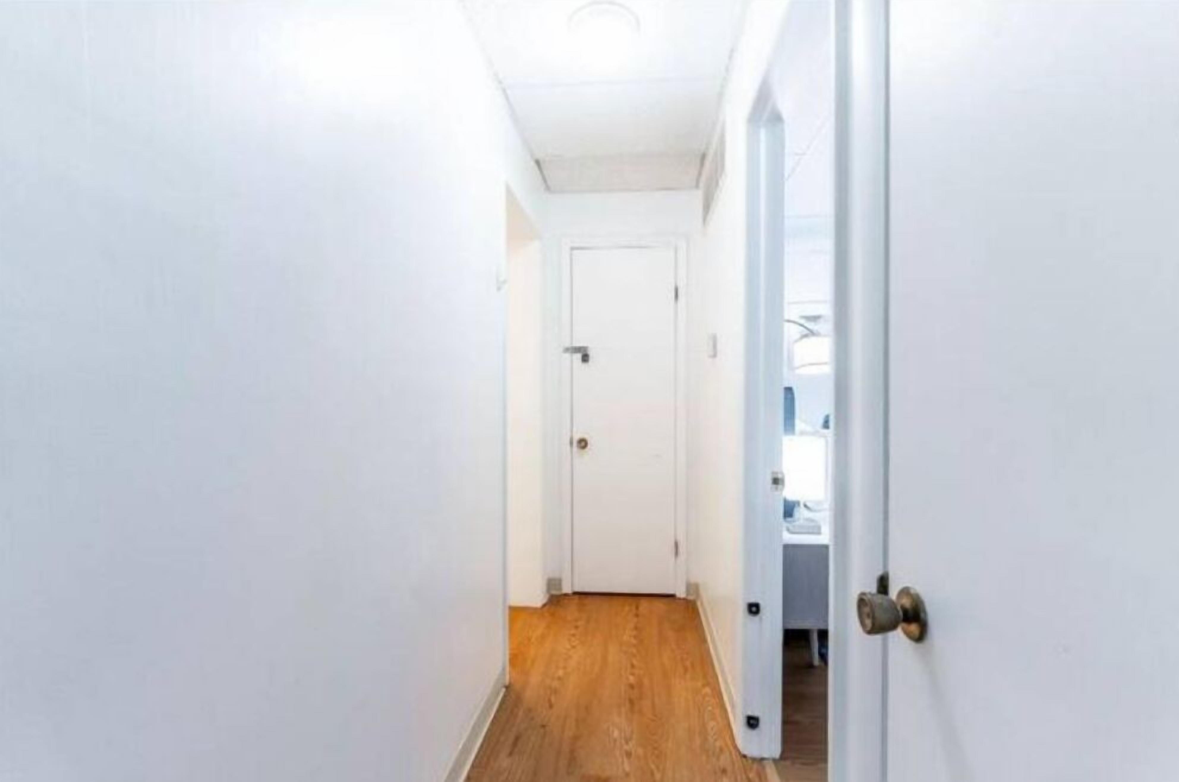 Un pasillo estrecho con paredes blancas y suelo de madera, que conduce a una puerta cerrada.