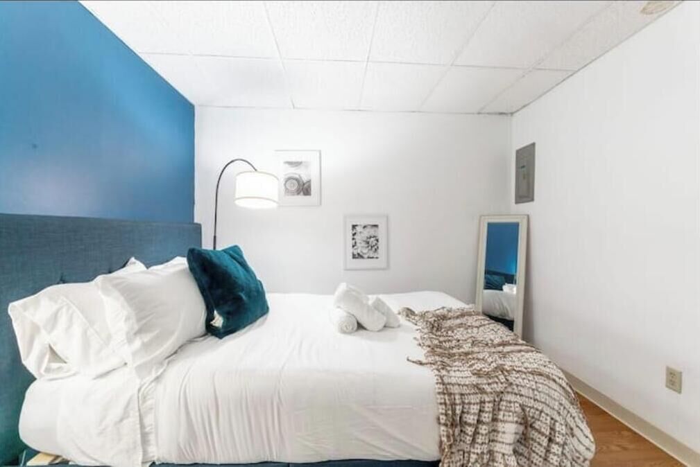 Un lit bien fait, avec du linge de lit blanc et un plaid à motifs, une lampe de chevet et des tableaux encadrés accrochés au mur.