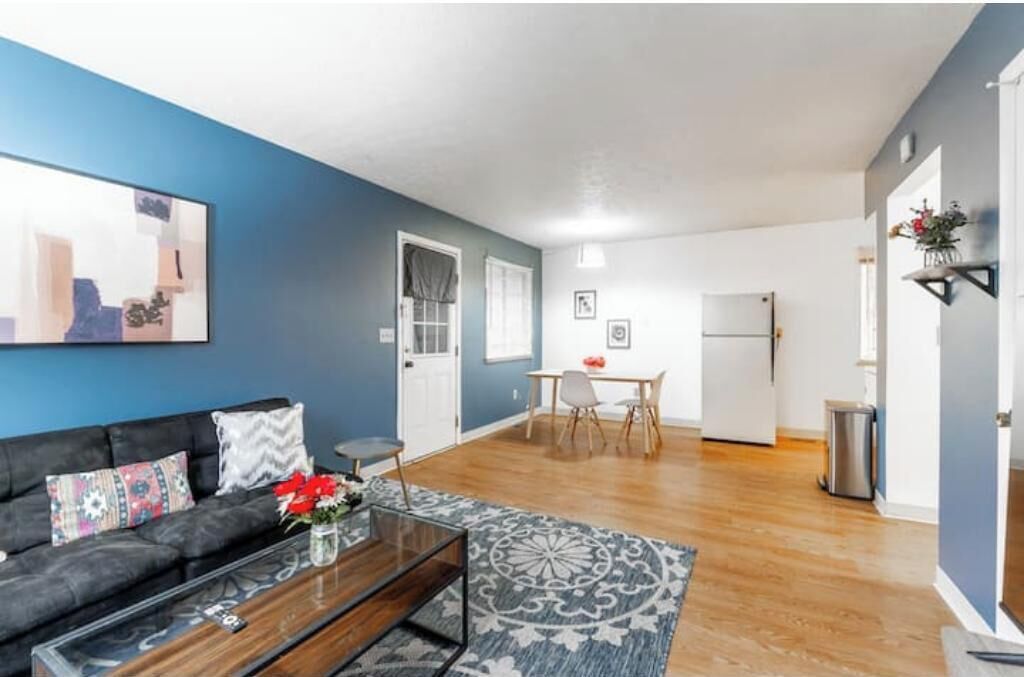 Un salon moderne avec un mur d’accent bleu, un canapé noir agrémenté de coussins à motifs, une table basse en verre et un coin repas avec une table et des chaises blanches.