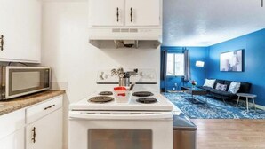 A compact kitchen with white cabinets, a microwave, and a stove. The room has a blue accent wall and a small seating area with a sofa and a coffee table. - OSU - Heart of Short North -STEPS from High ST-OSU (Columbus)