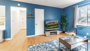 A living room with a blue wall, a wooden floor, a TV on a stand, a coffee table, and a window with curtains. - OSU - Heart of Short North -STEPS from High ST-OSU (Columbus)