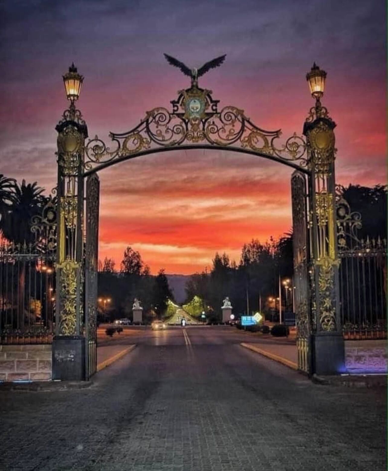 Un arco ornamentado con un emblema de águila, flanqueado por dos faroles, que conduce a una carretera con un atardecer en el fondo.