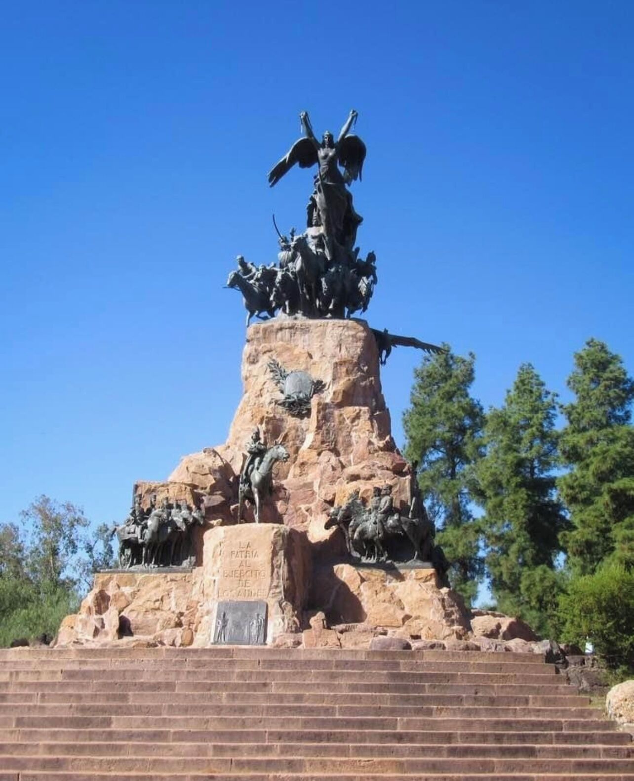 A large outdoor statue with multiple figures, including a central winged figure, on a rocky pedestal with steps leading up to it.
