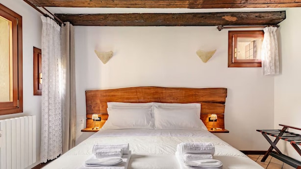 Una cama bien hecha con ropa de cama y toallas blancas, cabecera de madera, dos lámparas de pared y un ventanal con cortinas.