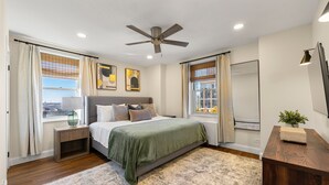 A bedroom with a bed, nightstand, bedside table, ceiling fan, window with curtains, and a view of the cityscape. - Stylish 1-bedroom condo Downtown Atlanta (Atlanta)
