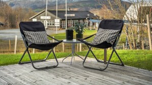 Deux fauteuils noirs confortables avec des coussins à carreaux, placés sur une terrasse en bois, une petite table avec un pot de plante, et une vue sur une montagne et des bâtiments en arrière-plan.