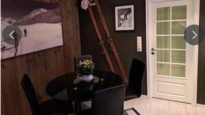 Un coin repas avec une table ronde, des chaises noires, une plante en pot, du matériel de ski et une porte dont les panneaux sont en verre dépoli.