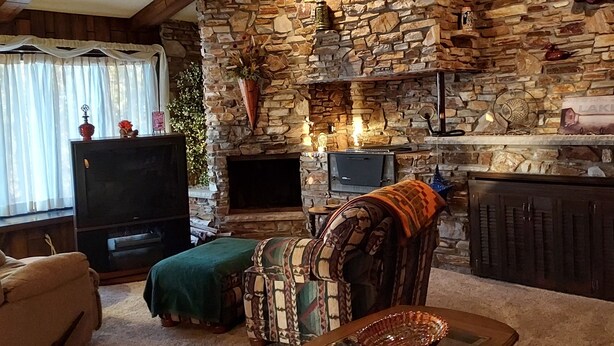 A cozy living room with a stone fireplace, a television, and a dining table with a glass top. - Charming 2-bedroom house in Shawano with AC, WiFi (Shawano)
