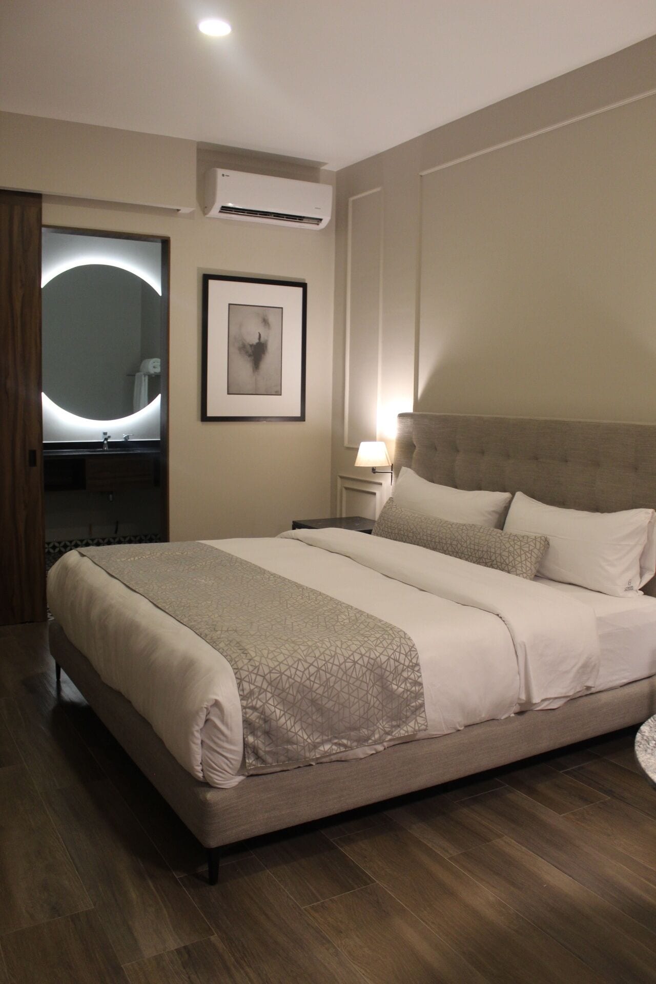 A neatly arranged bedroom with a large bed, a bedside table, a wall-mounted air conditioner, a framed abstract artwork, and a mirror with a circular light.