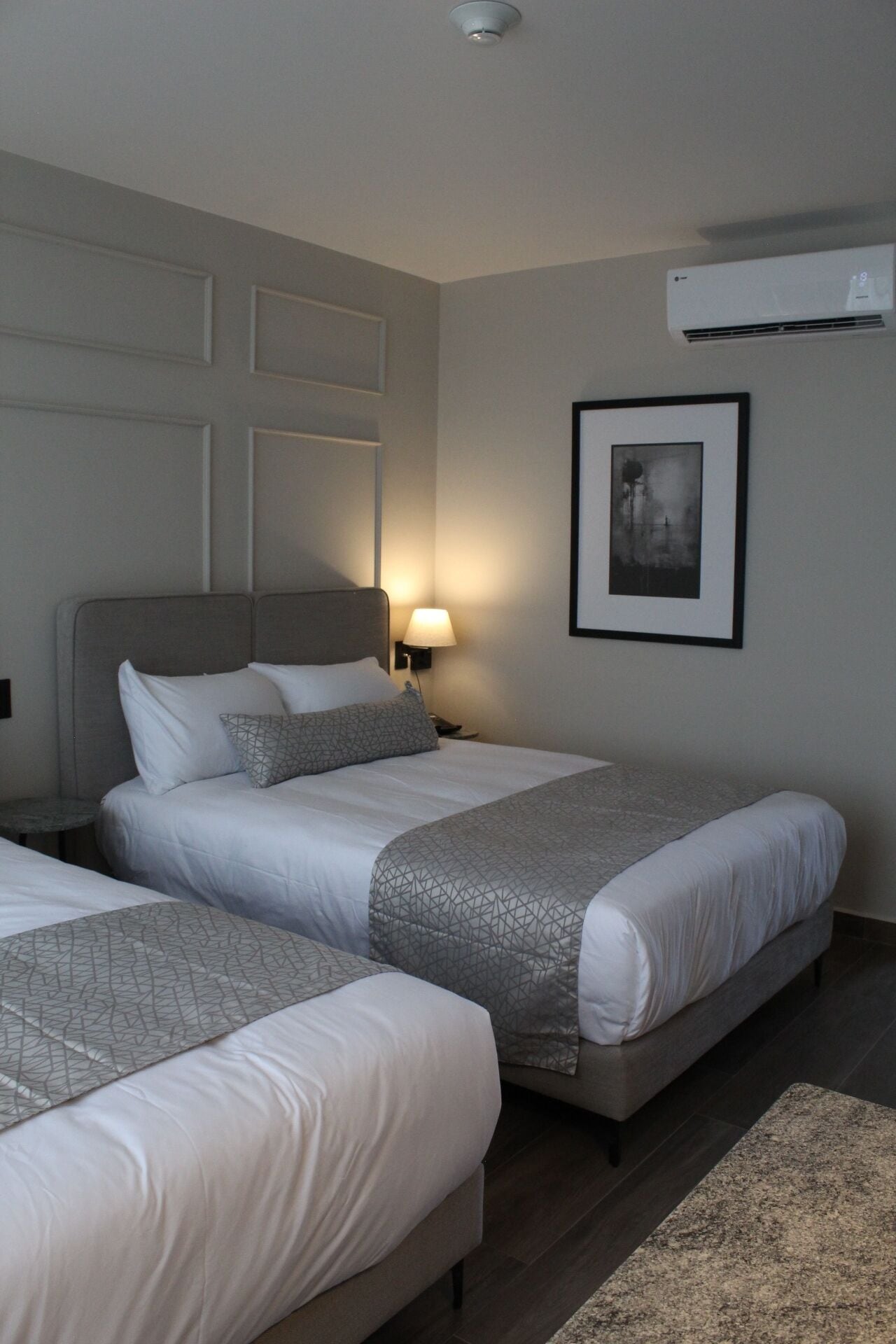 A hotel room with two beds, a bedside table with a lamp, a framed picture on the wall, and an air conditioning unit.