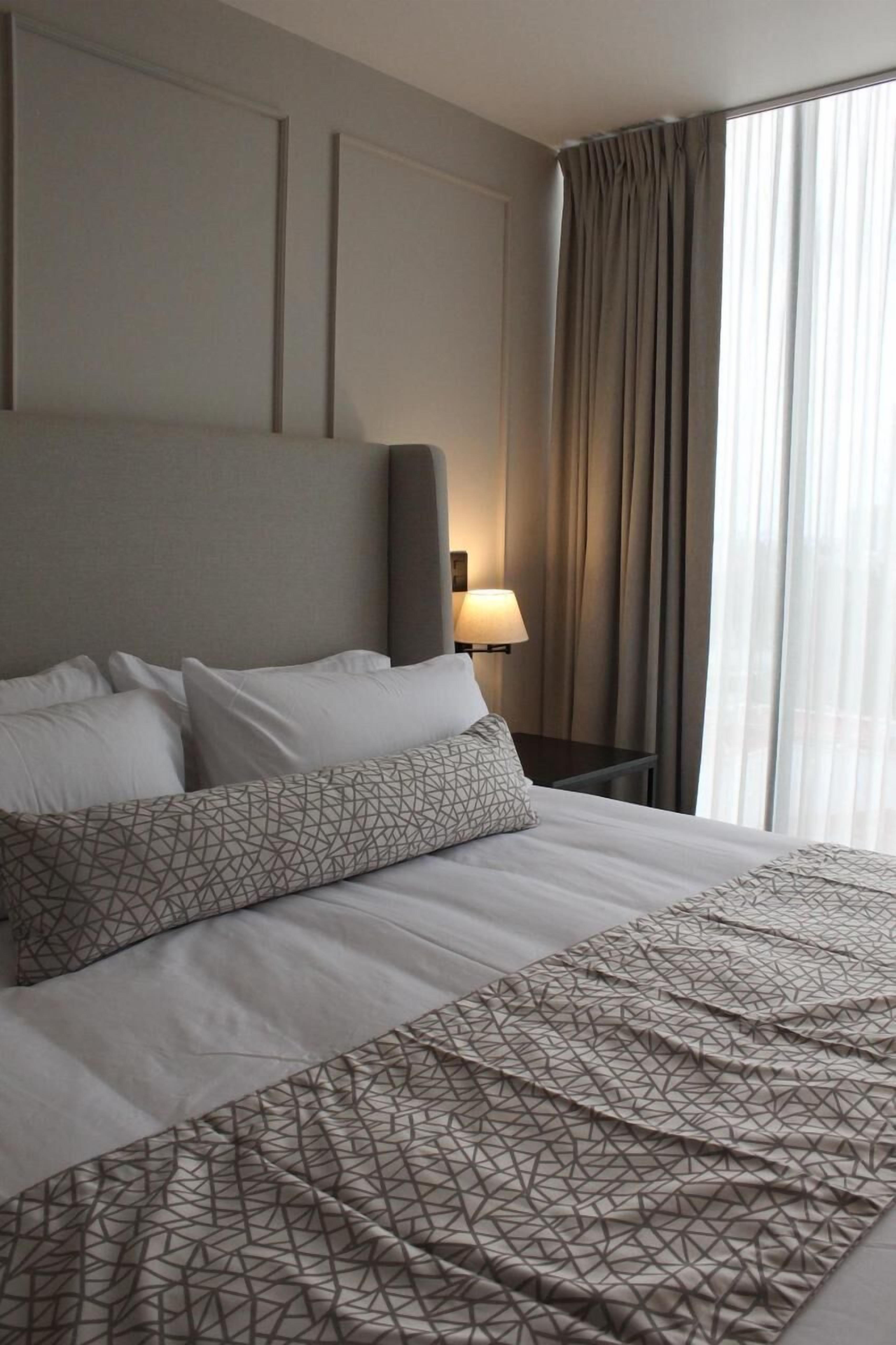 Una habitación de hotel con una cama grande, cabecera, mesita de noche con lámpara y ventana con cortinas.
