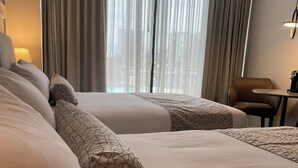 Habitación de hotel con dos camas, una silla, una mesa y vistas a la ciudad a través de amplios ventanales.