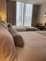 A hotel room with two beds, a chair, a table, and a view of the city through large windows.