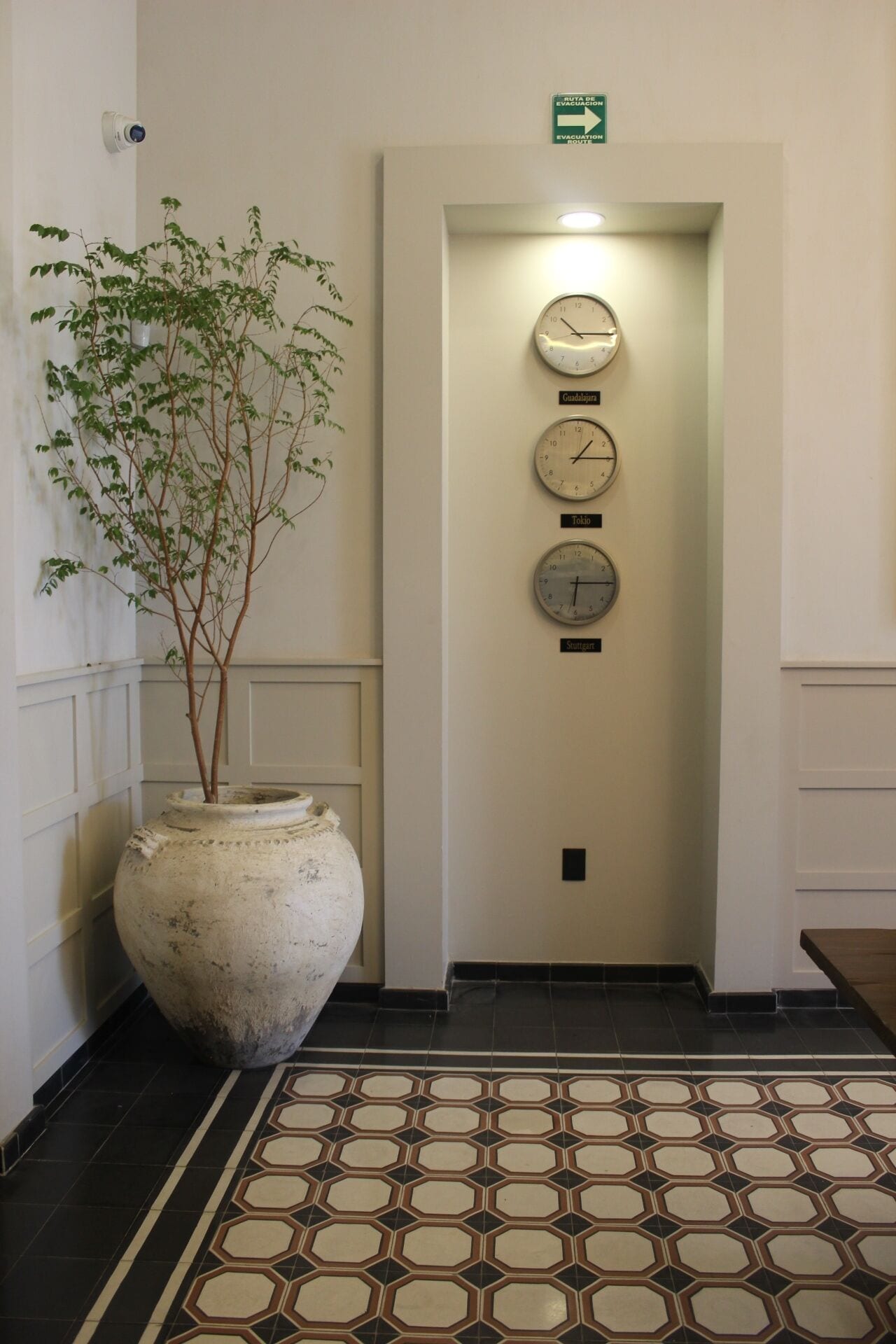 A hallway with a large vase, three clocks labeled 'Guadalajara,' 'Tokyo,' and 'Singapore,' and an emergency exit sign.