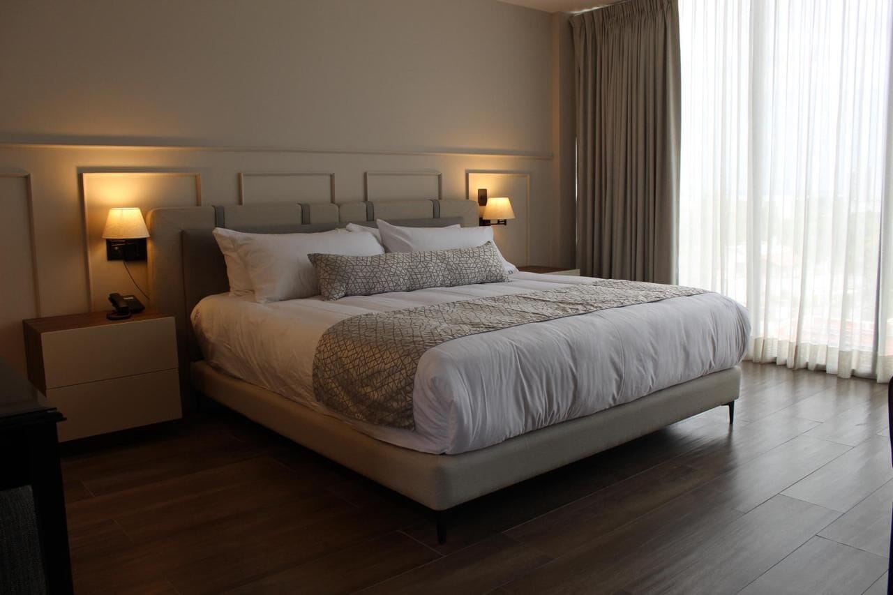 A modern hotel room with a large bed, two bedside tables with lamps, a wooden floor, and a view of the city through sheer curtains.