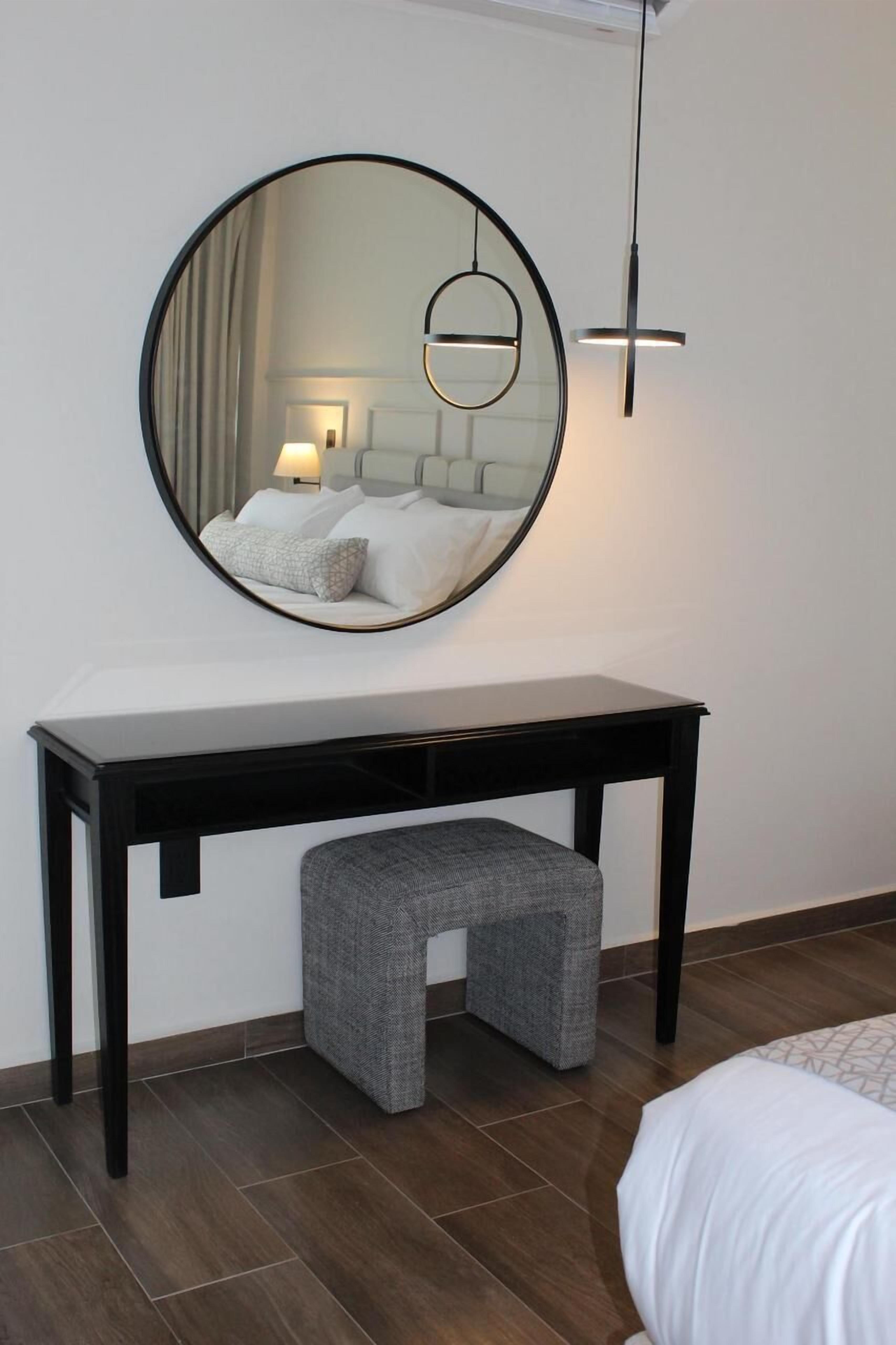A modern bedroom with a round mirror, a bedside table, a grey upholstered bench, and a bed with white bedding.