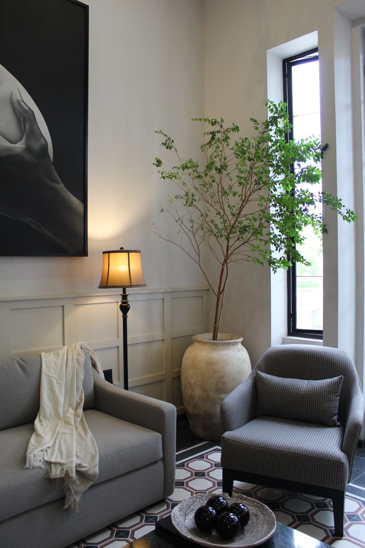 Ein Wohnzimmer mit einem grauen Sofa, einem gemusterten Sessel, einer großen Vase mit Pflanze, einer Stehlampe und einem Bild an der Wand.