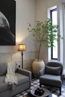 Una sala de estar con un sofá gris, un sillón estampado, un jarrón grande con una planta, una lámpara de pie y un cuadro en la pared.