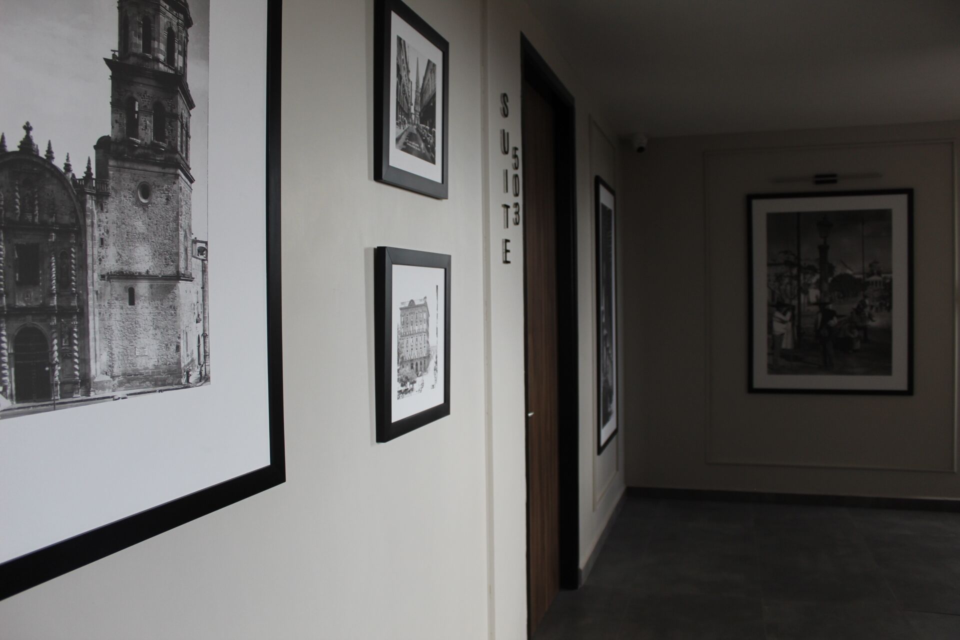 Un pasillo con fotografías en blanco y negro enmarcadas en las paredes, una puerta con la etiqueta “SUITE E” y una escalera que sube a otro piso.