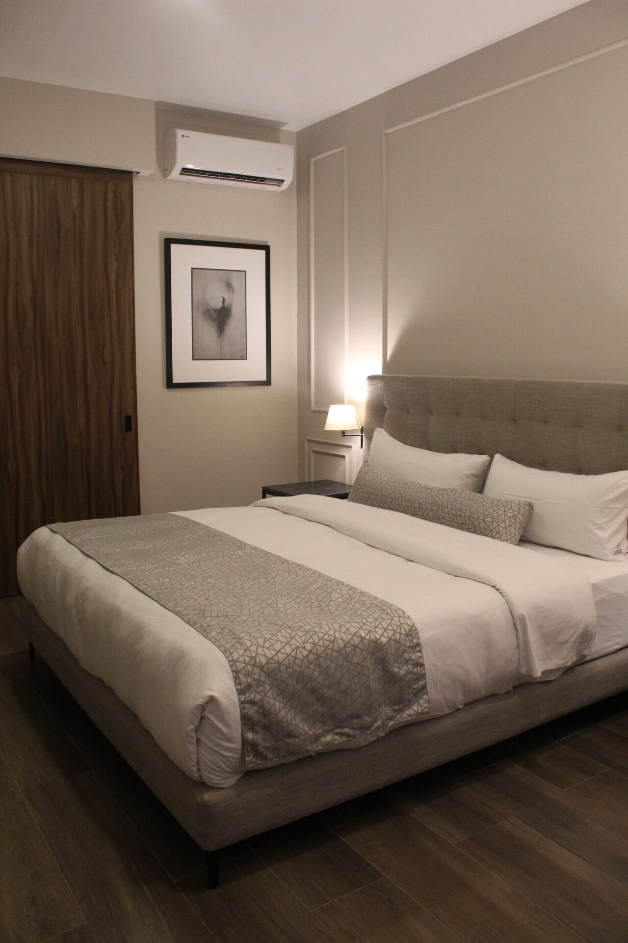 A neatly made bed with a geometric patterned bedspread, a framed abstract artwork on the wall, and a wall-mounted air conditioner.