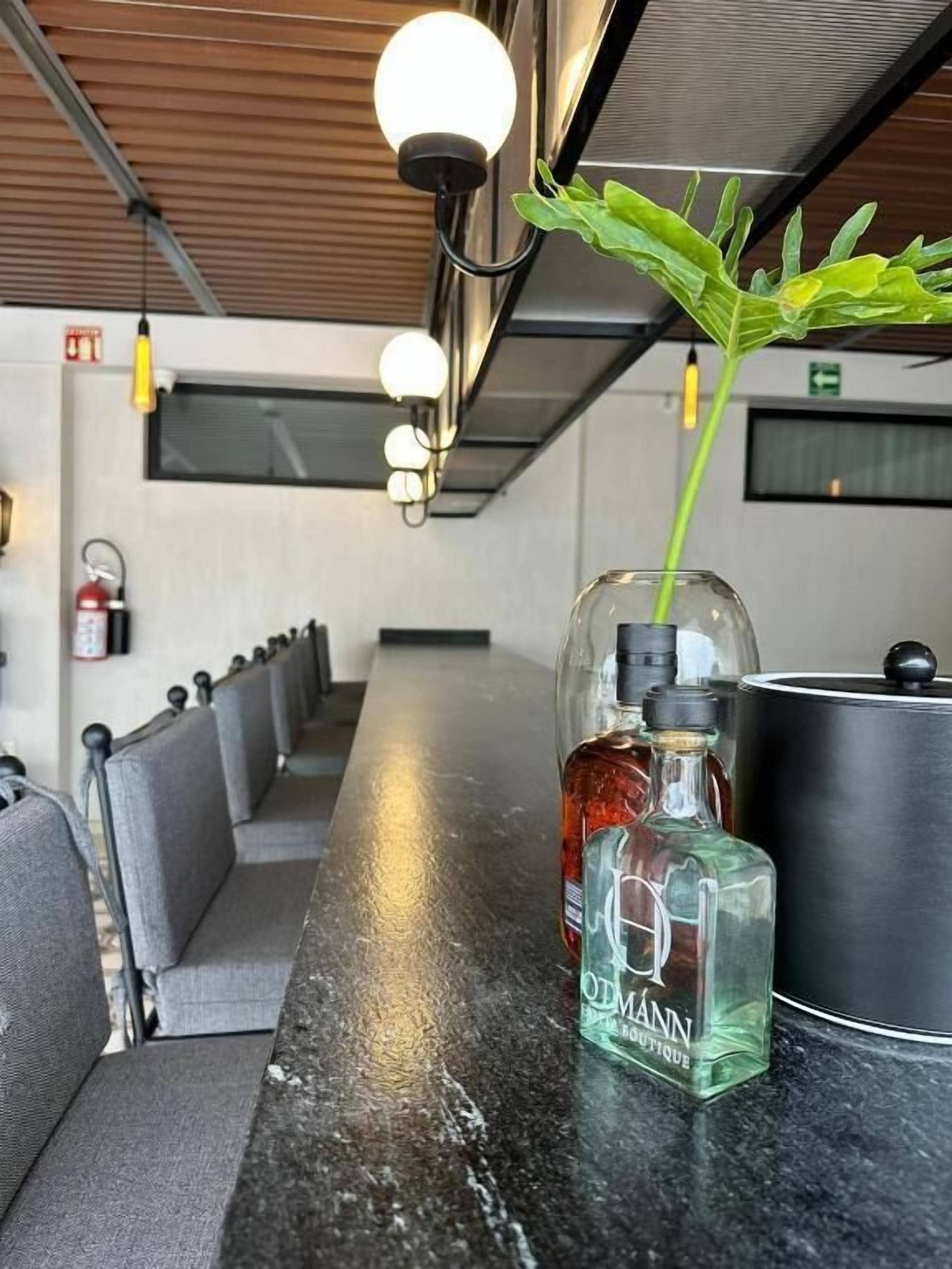 Una larga mesa de madera oscura con una botella de vidrio y un recipiente metálico encima. La mesa está rodeada por sillas tapizadas en gris.