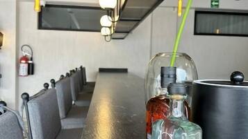 Une longue table en bois sombre, sur laquelle se trouvent une bouteille en verre et un récipient en métal. La table est bordée de chaises grises à revêtement rembourré.