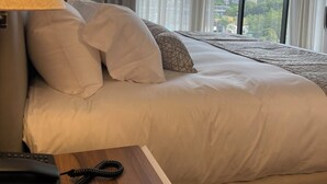 Habitación de hotel con una cama grande, una lámpara de noche, un teléfono y vistas de la ciudad a través de amplios ventanales.