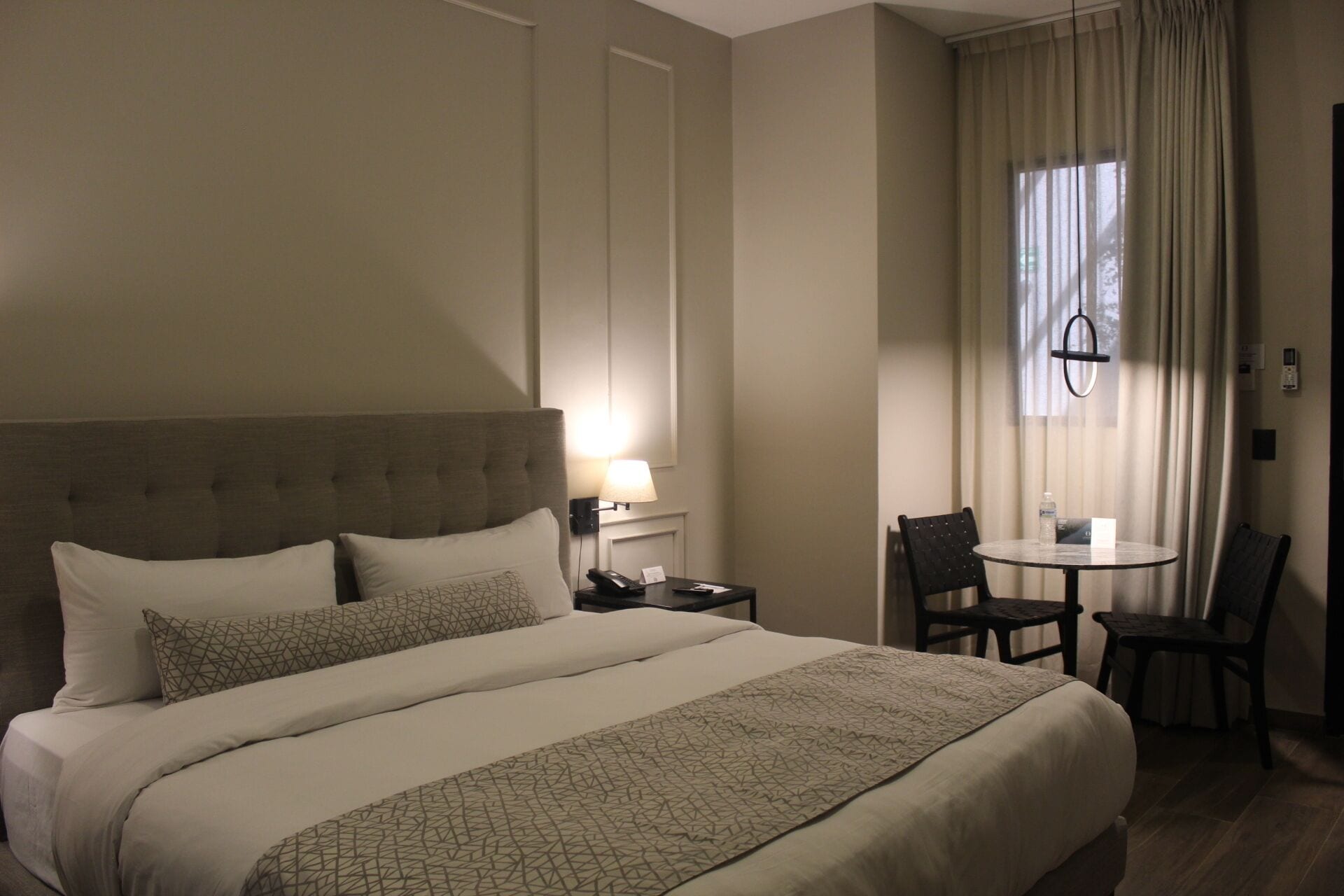 Hotellihuone, jossa on suuri sänky, yöpöytä lampun kanssa, pieni pöytä tuoleineen ja ikkuna, jossa on verhot.