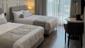 A hotel room with two beds, a desk, a chair, a coffee maker, and a large window with curtains.