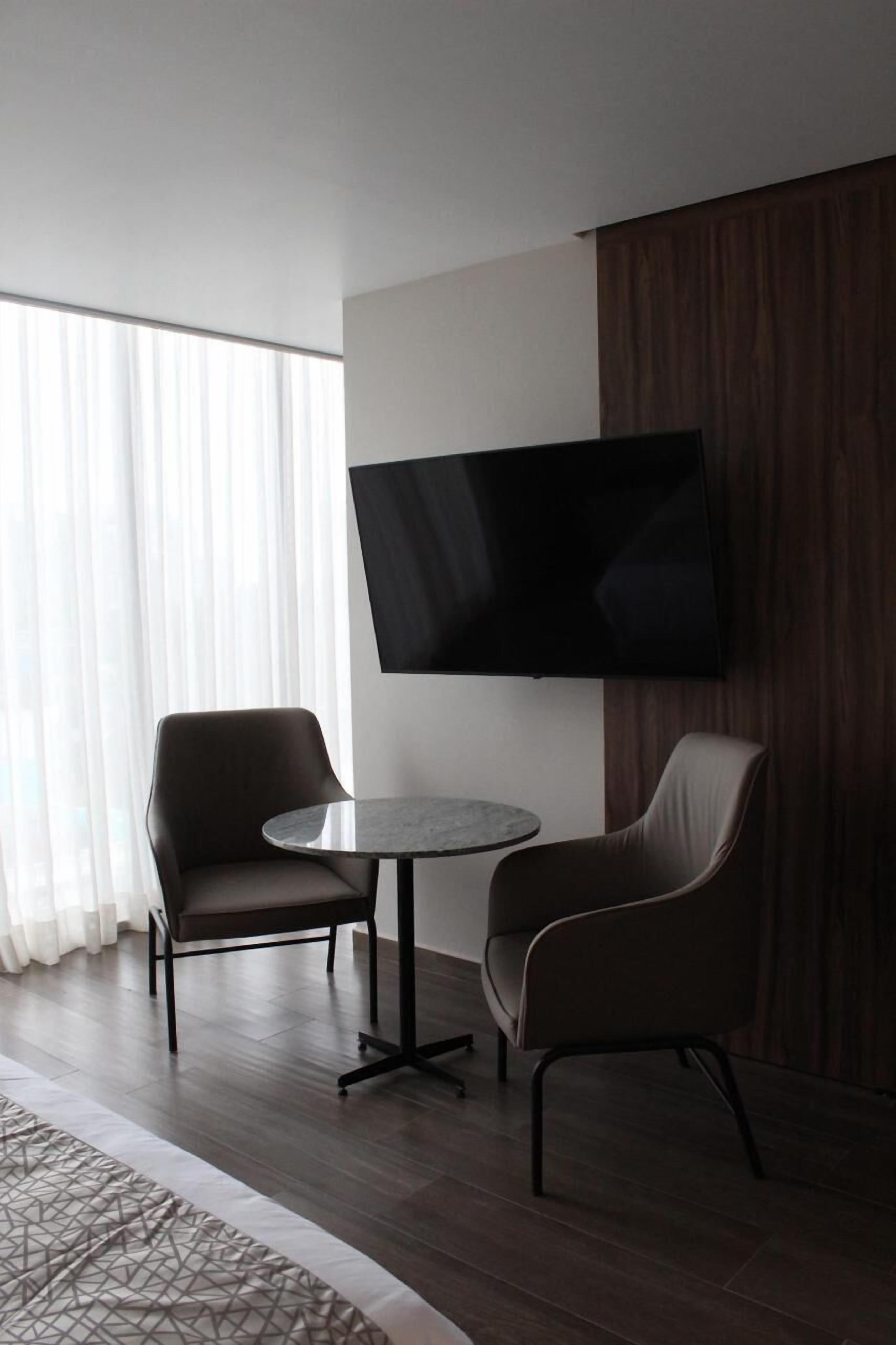 Een moderne hotelkamer met een grote flatscreen-televisie aan de muur, twee comfortabele fauteuils, een klein rond tafeltje en een groot raam met dunne gordijnen.