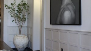 A hallway with a tiled floor, a large potted plant, a table with items, and a large framed portrait on the wall.