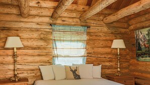 Un dormitorio tipo cabaña de madera con una cama grande, dos mesitas de noche con lámparas, una ventana con cortinas y un cuadro en la pared.