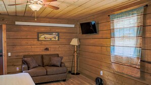 Una habitación acogedora con techo de madera, ventilador de techo, televisor de pantalla plana en la pared, sofá marrón con cojines, lámpara de pie y ventana con cortina a rayas.