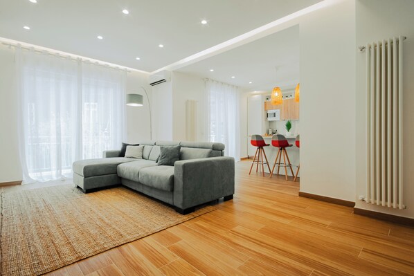 Un salon moderne avec un canapé d’angle gris, un plancher en bois et un coin cuisine à l’arrière-plan.