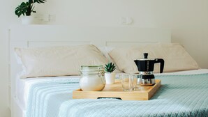 A neatly made bed with a light blue quilt, a wooden tray with a coffee maker, a glass jar, and a small plant.