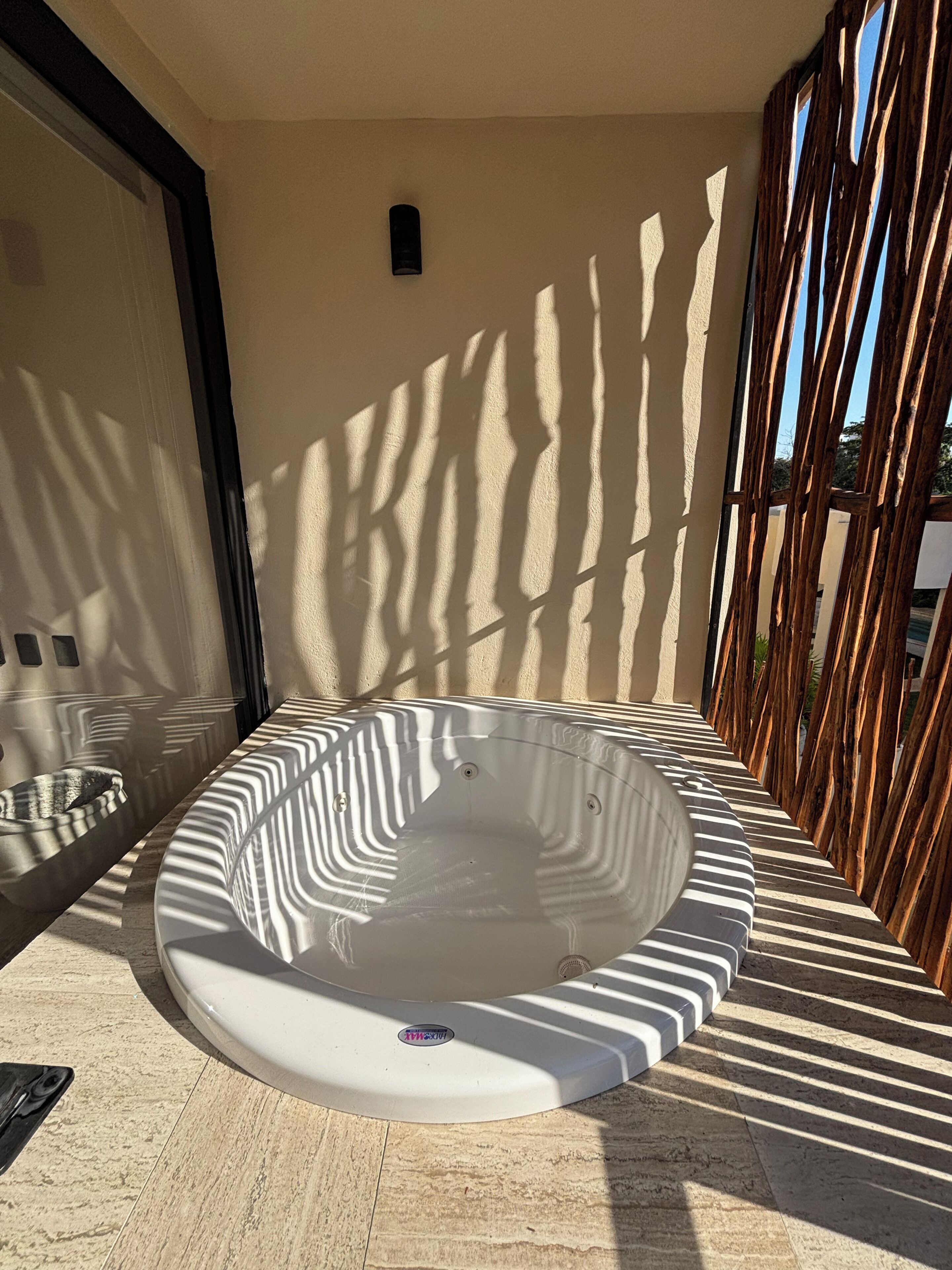 A hot tub in a sunlit outdoor area with a tiled floor, a metal bowl, and a wooden fence.