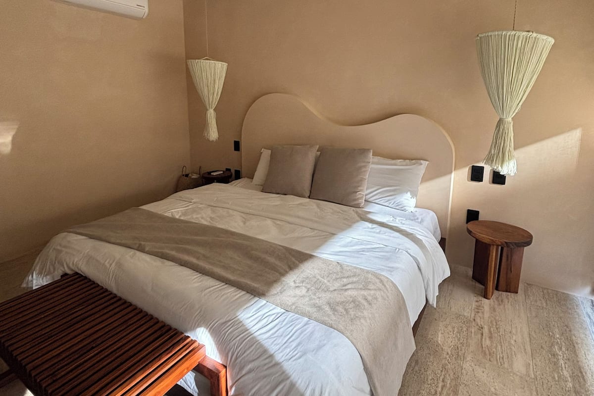 A bedroom with a bed, a ceiling fan, an air conditioning unit, a wooden bench, and two hanging lamps.