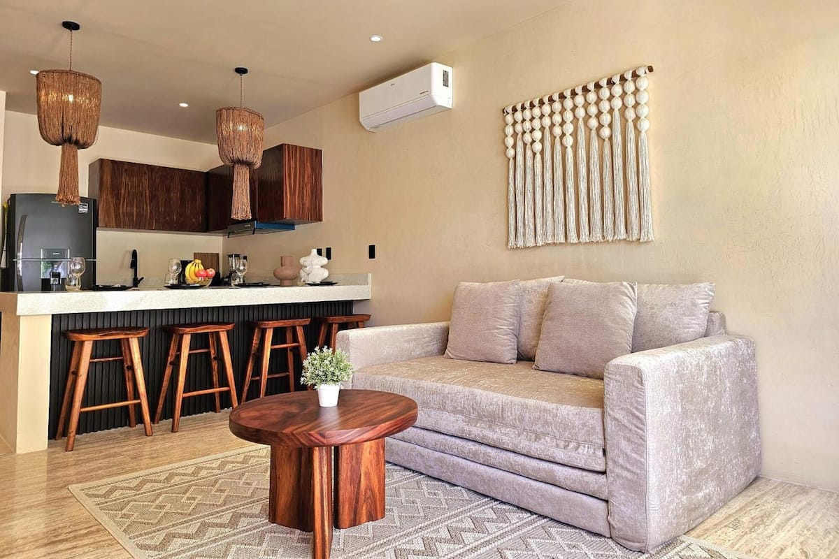 A modern living room with a sofa, a coffee table, and a kitchen area with stools and a refrigerator.