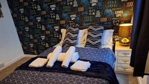 A neatly made bed with a geometric patterned bedspread and pillows, a bedside table with a lamp, and a wall with a repeating 'LEVEL UP!' pattern. - Fantastic Large Room with  free parking (Chatham)